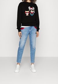 Felpa nera con personaggi dei cartoni animati colorati, abbinata a jeans azzurri chiari con accenti rossi e sneakers bianche. Design semplice.