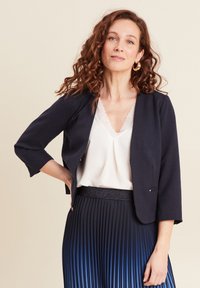 Giacca corta blu navy con maniche tre quarti, abbinata a una maglietta bianca con scollatura in pizzo e una gonna plissettata con sfumature di blu.