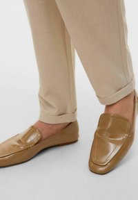 Beige leren instappers met een glanzende afwerking, voorzien van een gestikt detail op de wreef en een platte zool, gecombineerd met omgeslagen beige broeken.