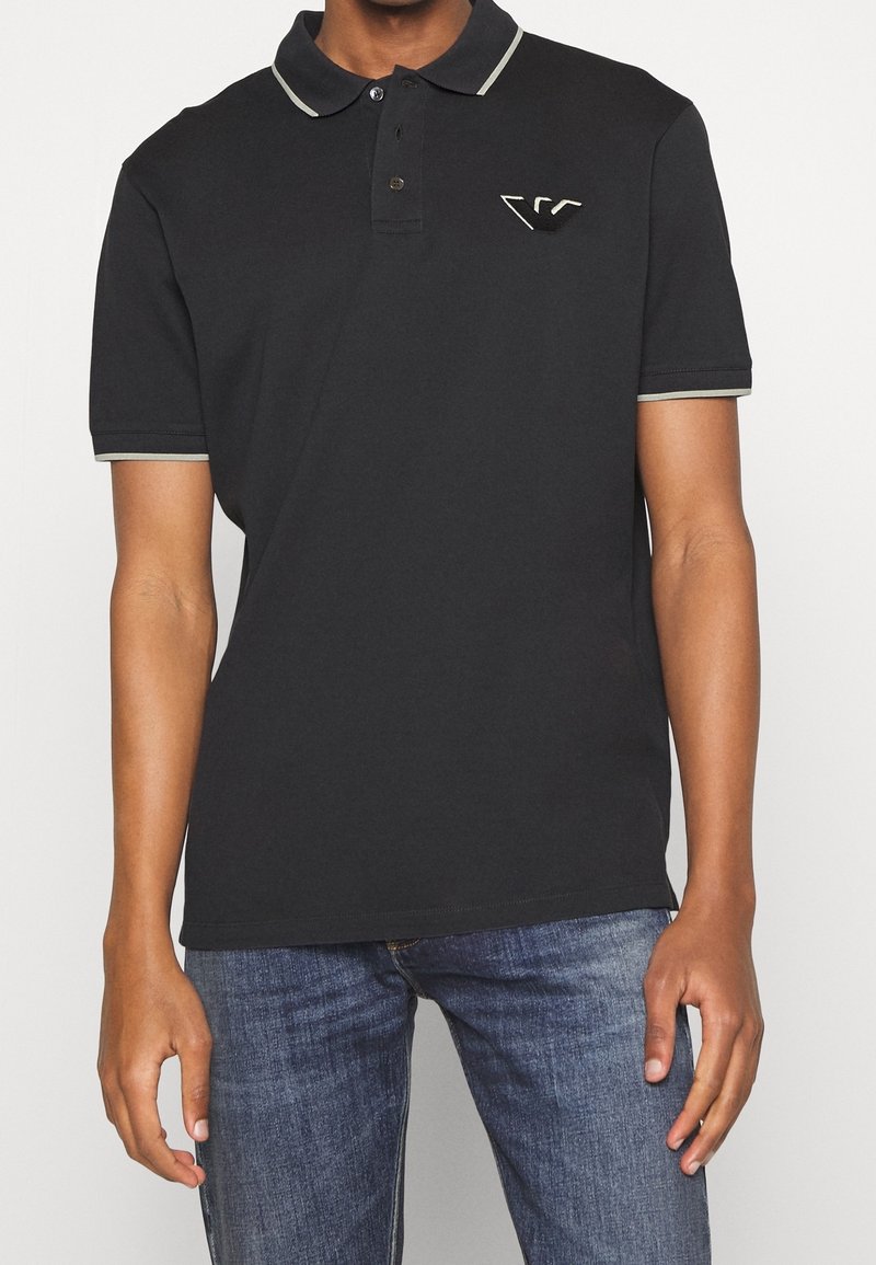 Homme portant un polo noir avec un liseré gris clair au niveau du col et des manches, associé à un jean bleu, debout devant un fond uni.
