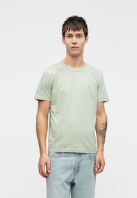 Light green cotton t-shirt with a crew neck, short sleeves, and a simple logo detail on the chest. Worn with light blue denim jeans.