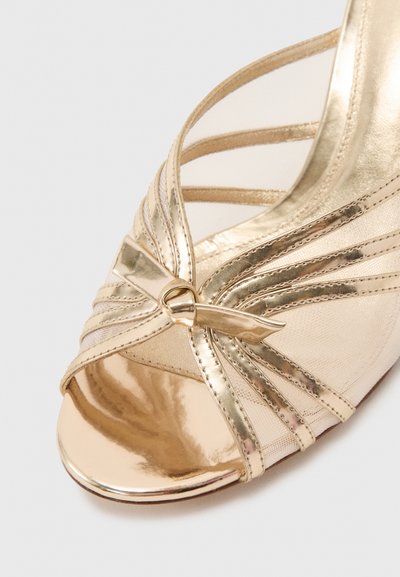 Χρυσό πέδιλο με ανοιχτό δάκτυλο και ψηλό τακούνι, διακοσμημένο με φιόγκο και πολλαπλές λεπτές λωρίδες σε ανοιχτό φόντο.