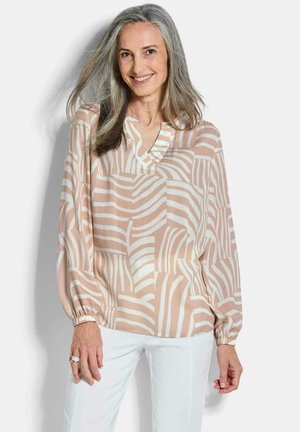 Lächelnde Frau mit langen grauen Haaren trägt eine beige-weiß gemusterte Bluse mit langen Ärmeln und weiße Hose vor schlichtem Hintergrund.