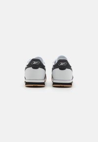 Chaussures de sport blanches et noires avec un bout arrondi, un col rembourré et une semelle en caoutchouc naturel, arborant un logo sur le talon et des détails texturés.