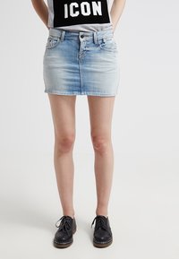 Person som bär en kort ljusblå jeanskjol, en vit och svart topp med texten "ICON", samt svarta snörskor, står på en ljus bakgrund.