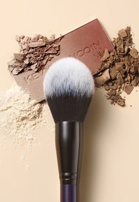 Make-up-Pinsel mit einem dichten, weiß-schwarzen Borstenkopf, neben einer Palette mit Puder in Beige-, Braun- und Bronzetönen, mit etwas verstreutem Puder.