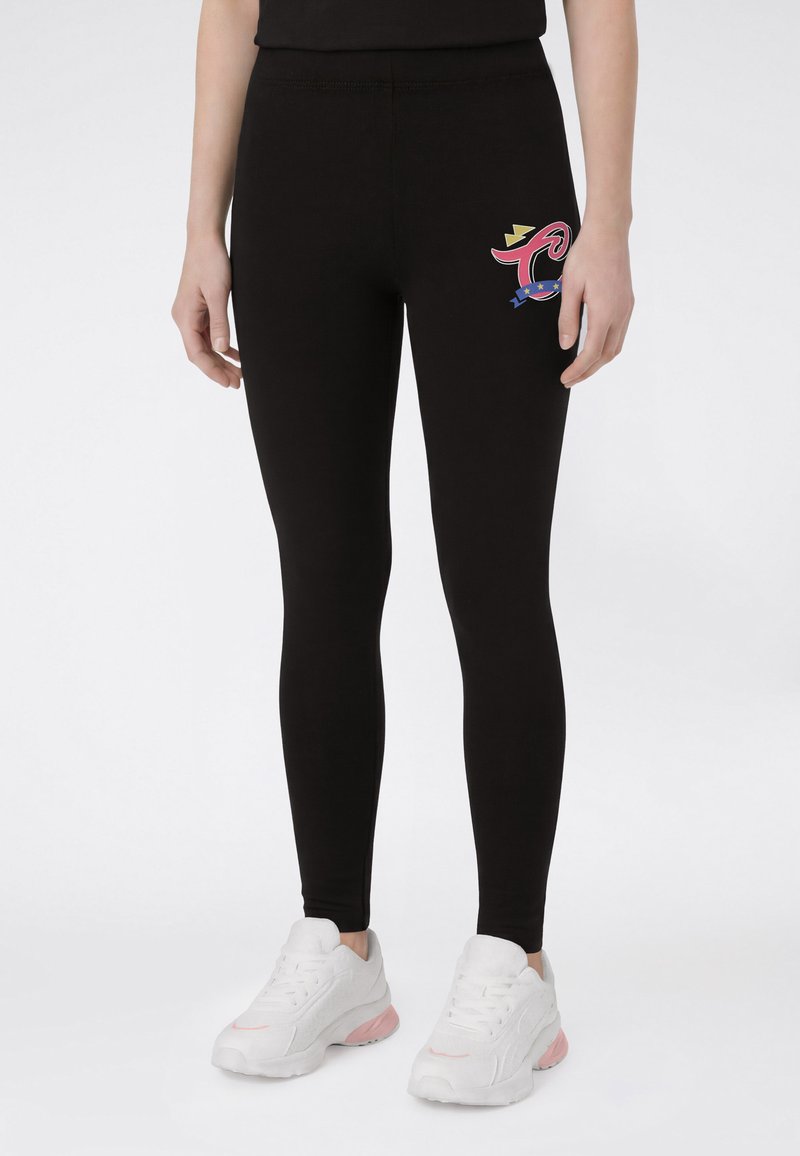 Personne portant un legging noir avec un motif coloré sur la cuisse gauche et des baskets blanches avec des semelles roses, debout sur une surface blanche.
