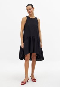 Musta ilmanhihaisilla mekko, jossa on teksturoitu pinta, rypytetty helma ja korkea-matala muotoilu. Yhdistetty punaisiin avokkaisiin.