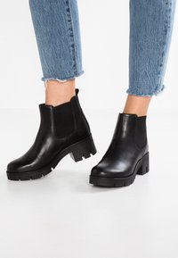 Botas negras de cuero hasta el tobillo con paneles laterales elásticos, suelas gruesas y un acabado suave, combinadas con jeans azules doblados.