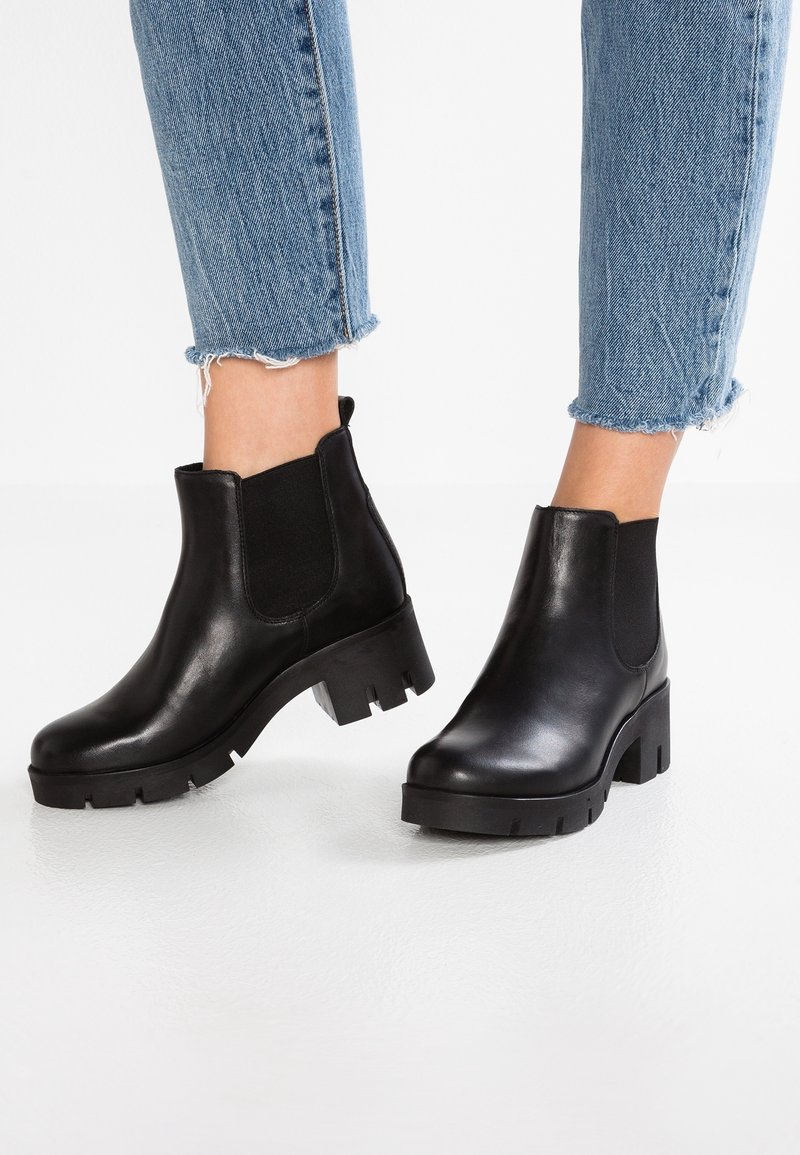 Botas negras de cuero hasta el tobillo con paneles laterales elásticos, suelas gruesas y un acabado suave, combinadas con jeans azules doblados.