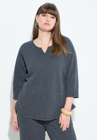 Donna con lunghi capelli castani che indossa un abbigliamento coordinato di top e pantaloni blu scuro con texture, in piedi contro uno sfondo bianco, con un braccialetto beige al polso.
