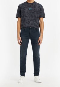 Camiseta de hombre de Levi's en gris oscuro con un patrón de nubes y un logo azul, combinada con jeans de mezclilla oscuros y zapatillas blancas.