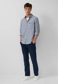 Camicia azzurro chiaro, con bottoni e tasca sul petto, maniche arrotolate e tessuto strutturato. Abbinata a pantaloni blu navy e sneakers bianche.