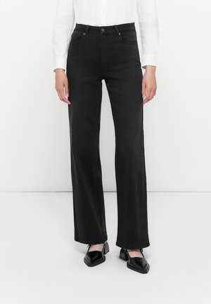 Woman wearing high-waist black straight-leg jeans, white long-sleeve shirt, and black square-toe heeled shoes standing on white floor.