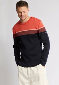 Homme portant un pull rouge et marine à rayures horizontales avec des boutons à l'épaule, associé à un pantalon clair, se tenant les mains dans les poches.