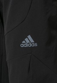 Vêtement de sport noir en matériau extérieur avec un design géométrique, arborant un logo Adidas gris clair bien en vue.