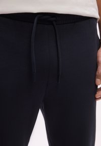 Κοντινό πλάνο σκούρων μπλε φόρμας με κορδόνι στη μέση, που φοριέται από άτομο με λευκό πουκάμισο, εστιάζοντας στη μπροστινή δέση και τη χαλαρή εφαρμογή.