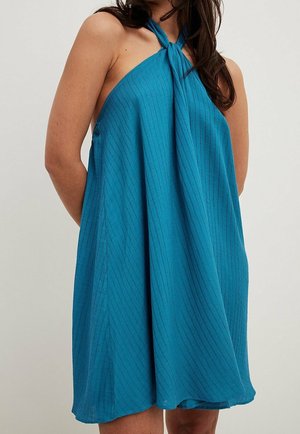 Vrouw draagt een mouwloze, blauwe textuur halterjurk met gekruiste bandjes over blote schouders.