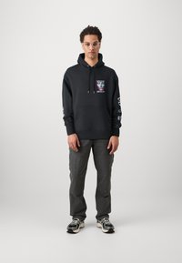 Sweatshirt preto com um gráfico na frente. Apresenta um bolso canguru, punhos e bainha canelados. Combinado com calças cargo cinzas e sapatilhas desportivas.