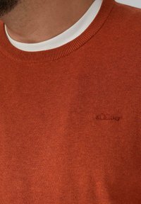 Close-up van een man die een roest-oranje gebreide trui draagt over een witte crewneck shirt, met een klein geborduurd s.Oliver logo.
