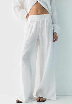 Személy laza, fehér, texturált, bő szárú nadrágot és hozzá illő, fehér hosszú ujjú felsőt visel, mezítláb áll fehér padlón.