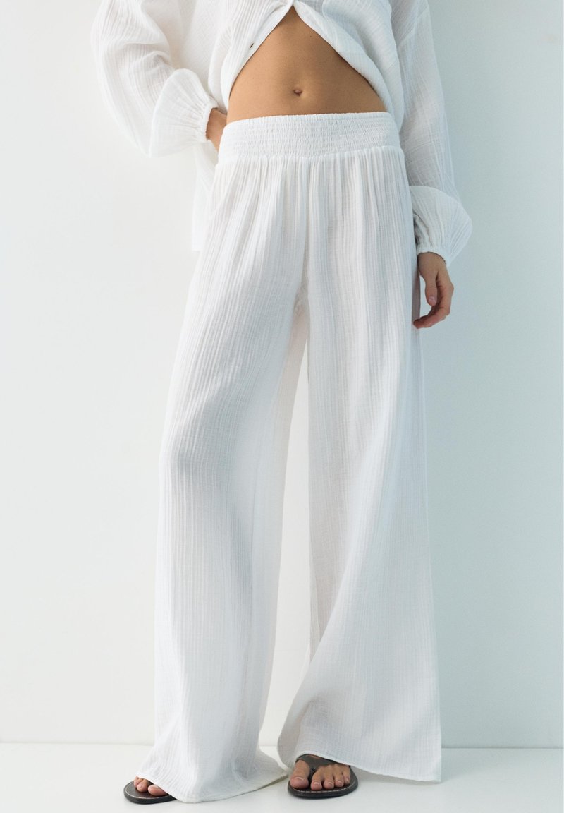 Personne portant un pantalon large blanc texturé et ample, avec un haut blanc à manches longues assorti, debout pieds nus sur un sol blanc.