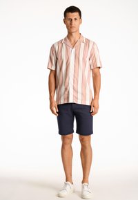 Camicia a maniche corte con bottoni, a righe verticali rosa e bianche, abbinata a pantaloni corta blu e sneakers bianche. Tessuto dalla texture liscia.