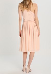 Robe sans manches de couleur pêche avec un corsage ajusté et une jupe froncée de longueur genou. Tissu lisse avec des détails de couture subtils à la taille.