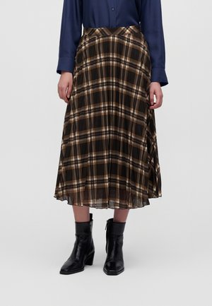 Femme portant une jupe midi plissée à carreaux noir, marron et blanc, une chemise à manches longues bleu marine et des bottines noires à talons carrés.