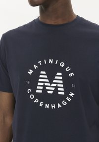 Tamsiai mėlynas medvilninis marškinėliai, kuriuose yra apvalus baltas logotipas su tekstu "MATINIQUE COPENHAGEN" ir dryžuotą raides "M" centre.