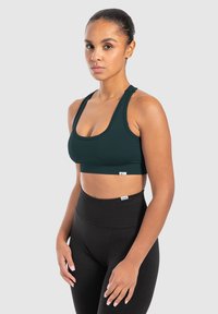 Tummanvihreä urheil bra, jossa on leveät hihnat, pyöreä kaula-aukko ja pehmeä vyötärö. Yhdistetty korkeavyötäröisten mustien legginsien kanssa.