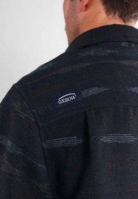 Hombre que lleva una chaqueta de patrón oscuro con una etiqueta de marca OXBOW visible en la parte superior de la espalda cerca del cuello.