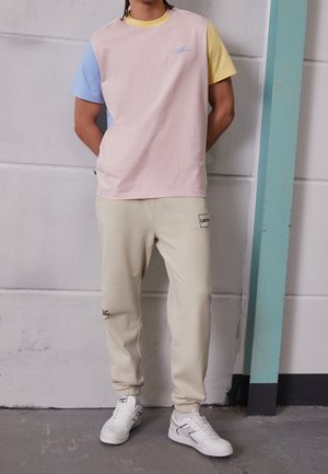 Person iført en pastelfarvet t-shirt med korte ærmer, beige joggerbukser med logoer og hvide sneakers, der står mod en betonvæg.