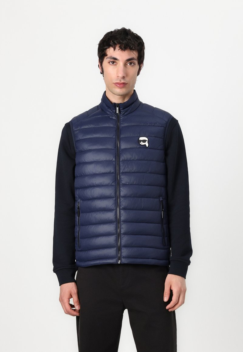 Marineblauwe gewatteerde vest met een hoge kraag, ritsluiting en twee zijzakken. Heeft een klein logo op de linkerkant van de borst; soepele textuur.