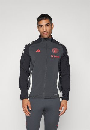Giacca a vento nera Adidas con logo rosso del Manchester United. Presenta strisce laterali bianche e una zip completa. Realizzata in materiale leggero.
