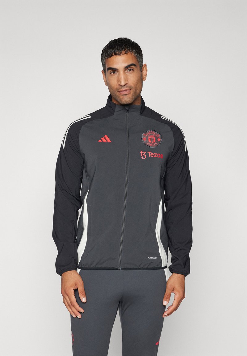 Chaqueta cortavientos negra de Adidas con el logo rojo del Manchester United. Cuenta con franjas laterales blancas y un cierre completo. Está hecha de material ligero.