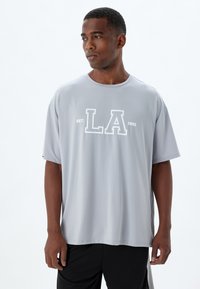 Camiseta gris de manga corta fabricada con un tejido suave, que presenta un gráfico blanco prominente que dice "LA" y "EST: 1992" en el frente.
