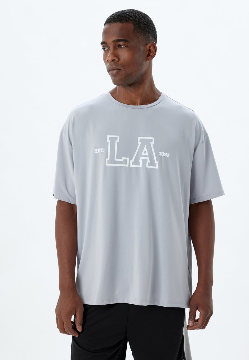 Camiseta gris de manga corta fabricada con un tejido suave, que presenta un gráfico blanco prominente que dice "LA" y "EST: 1992" en el frente.