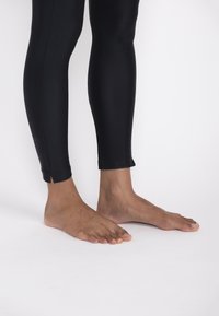 Black leggings with a smooth, stretchy texture tapering to the ankles, featuring small slits. Bare feet are visible at the bottom.