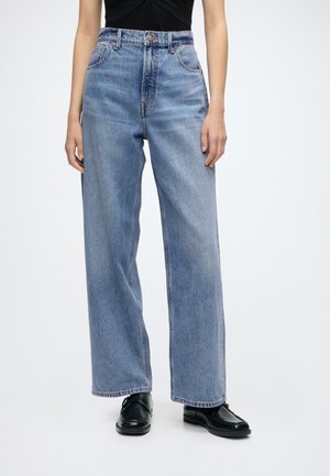 Femme portant un jean taille haute bleu clair à jambes larges avec poches avant, associé à des mocassins noirs et un haut noir rentré.