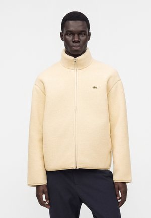Homme portant une veste polaire beige zippée avec un col haut et un pantalon sombre, debout devant un fond blanc uni.