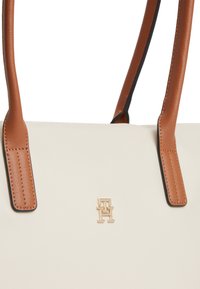 Τσάντα τότε κρεμ με καφέ δερμάτινα χερούλια και χρυσό διαπλεγμένο λογότυπο "TH" στο κέντρο της μπροστινής πλευράς.