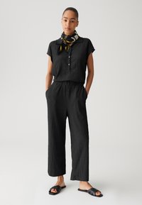 Zwart gestructureerd shirt met korte mouwen en knopen, gecombineerd met losse zwarte broek en platte zwarte sandalen. Een gestreepte sjaal accentueert de look.