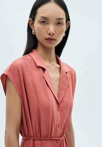 Abito camicia rosa pallido con maniche corte, colletto e chiusura con bottoni frontale. Il tessuto sembra leggero con un dettaglio di vita arricciata.