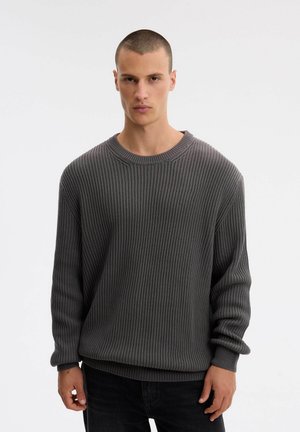 Un modelo masculino está de pie frente a un fondo liso y claro, con un jersey gris oscuro de canalé y cuello redondo y unos pantalones negros.
