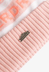 Różowa czapka beanie o prążkowanej strukturze z białym i pomarańczowym paskiem oraz metalową metką z logo. Miękka w dotyku, z wyraźnym kontrastem kolorów i wzorem.