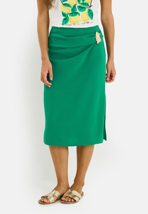 Mujer con falda midi verde con drapeado lateral y broche dorado, combinada con un top blanco con estampado de limón y sandalias planas doradas.