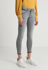 Šedé úzké džíny s střední vysokým pasem, které mají klasický pětikusový design, vybledlou texturu a roztrhaný lem, nošené s bílými teniskami.