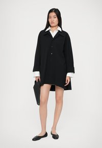 Černé oversized košilové šaty s knoflíkovým zapínáním, velkým límcem a bílými manžetami, doplněné o černé baleríny a černý desky.