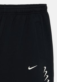 Črne trenirke z elastičnim pasom, stranskimi žepi in belim logotipom Nike. Imajo teksturiran zaključek in poenostavljeno obliko.